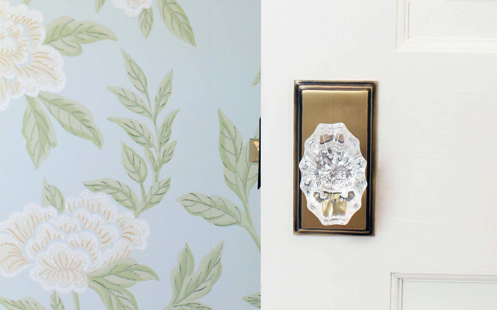 a white bedroom door featuring grandeur hardware vintage brass carre short plate with crystal knob with floral blue wallpaper in the background